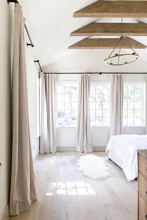 an airy and neutral bedroom with French windows, wooden beams, neutral furniture, pale pink curtains is ethereal