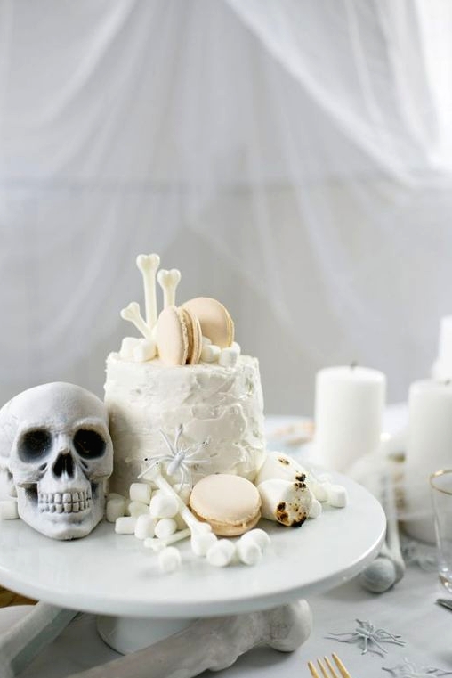 a white Halloween cake with edible bones, a skull, marshmallows and macarons is an adorable idea
