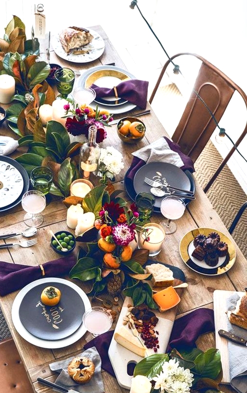 a bright Thanksgiving table setting with an uncovered table, a greenery and fruit runner, candles, blooms and black chargers