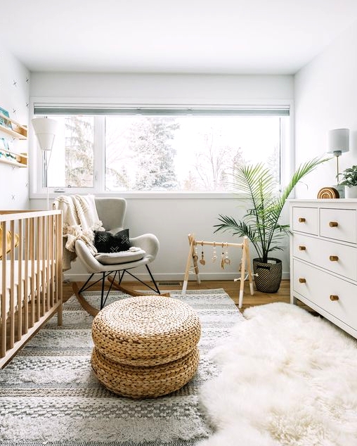 a fresh and inviting Scandi nursery with a wallpaper wall, a large stained crib, a grey rocker, a white dresser, layered rugs, open shelves