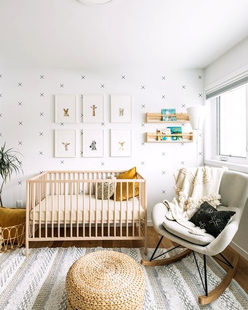 a neutral nursery with a wallpaper accent wall, a light stained crib, jute poufs, a white rocker, a grid gallery wall and open shelves