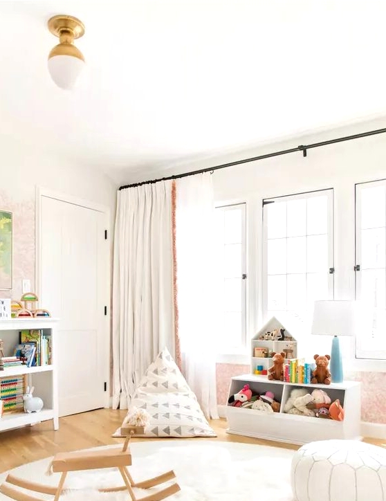 a pretty Scandi nursery with white furniture, white layered rugs, some touys, poufs and neutral curtains and pastel touches