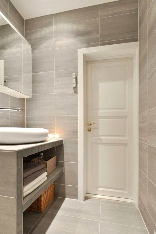 a modern bathroom clad with greige large scale tiles, with a vanity clad with them, too, a catchy white sink and a mirror cabinet over it