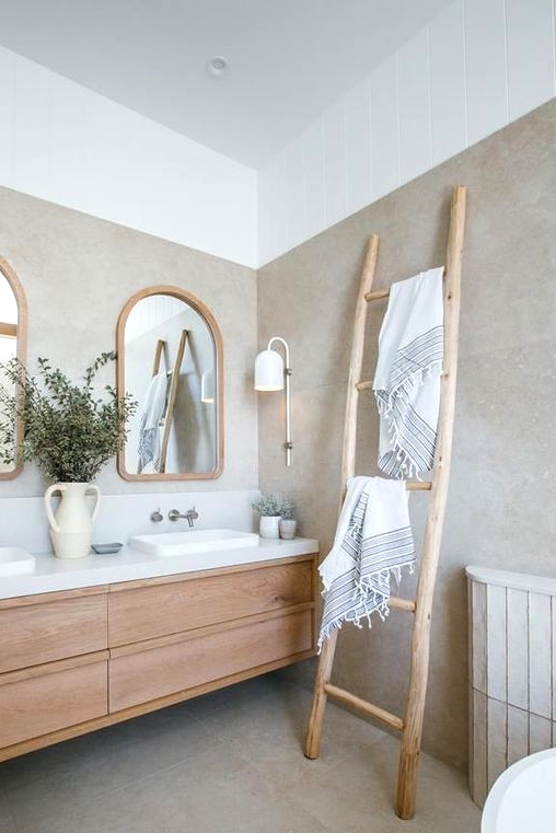 a serene and welcoming greige bathroom with a light-stained floating vanity, a ladder, arched windows in wooden frames and a cool sconce