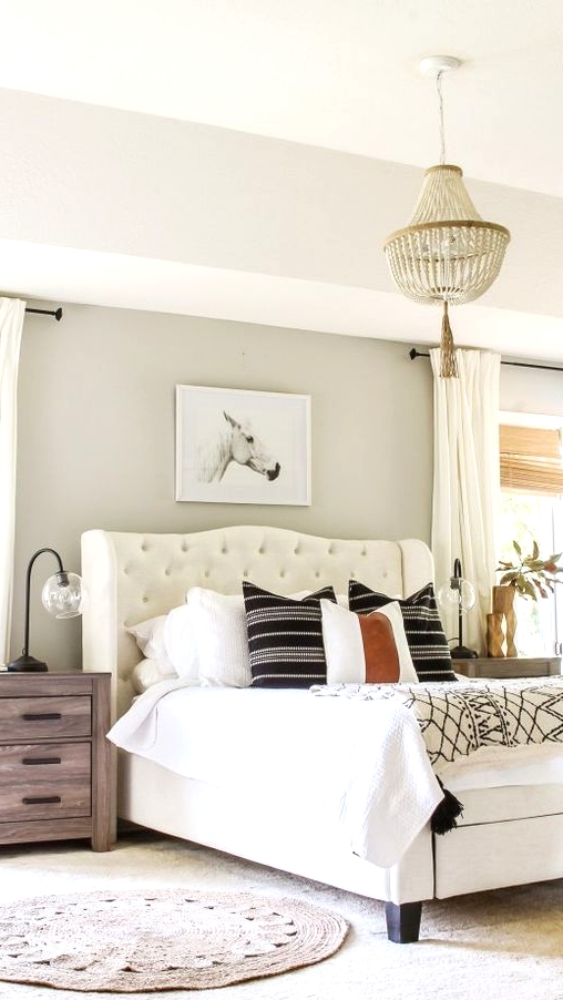 a cozy farmhouse bedroom with greige walls, a creamy upholstered bed, stained nightstands, neutral and printed bedding, a wooden bed chandelier and neutral curtains