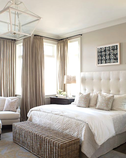 a welcoming greige bedroom with a creamy bed with a statement upholstered headboard, creamy seating furniture, a woven chest bench, greige curtains and a pendant lamp