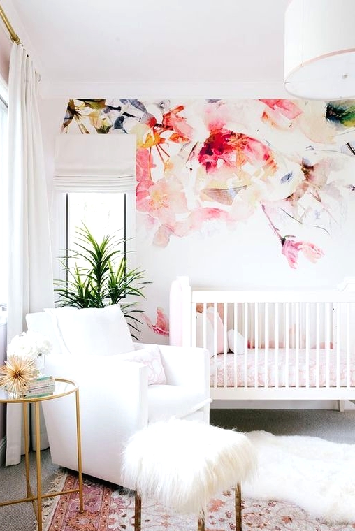 a bright contemporary nursery with a watercolor flower accent wall, a white chair and a crib, pink bedding and textiles, a little gold side table