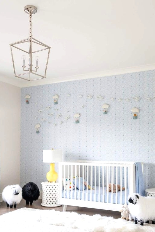 a light blue contemporary nursery with a blue accent wall with air balloons, a white crib and side tales, layered rugs and sheep toys