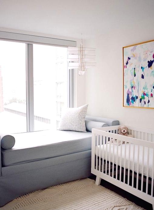 a simple contemporary nursery with a white crib, a colorful artwork, a grey daybed, a neutral rug and a view of the city