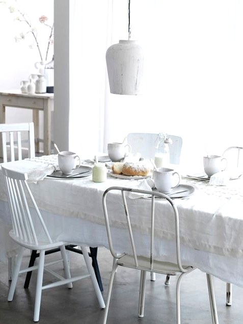 an all-white and serene Scandinavian dining room with a table covered with a white tablecloth, mismatching white chairs and a white pendant lamps is super inviting
