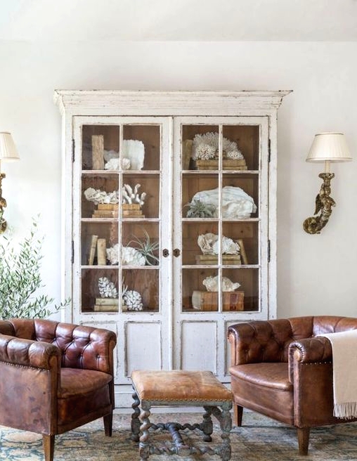 a shabby chic space with a whitewashed display cabinet with glass doors and leather chairs, a leather sotol and wall sconces