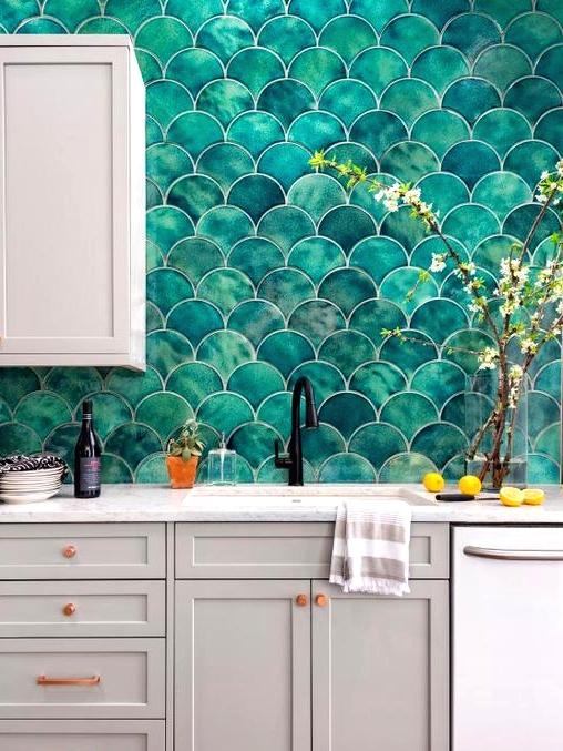 a grey kitchen with shaker cabinets, white stone countertops, a bold green fishscale tile backsplash and copper handles