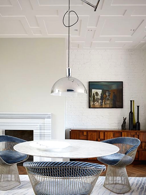 a beautiful eclectic dining room with white brick walls, a greige touch, a fireplace, a round table, blue chairs, a shiny pendant lamp and vases