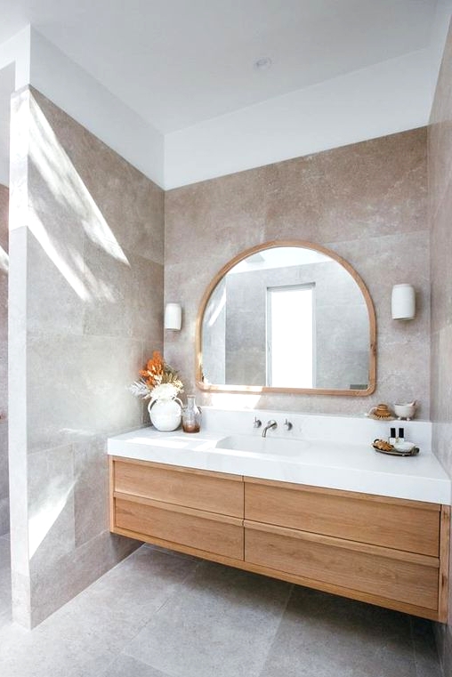 a beautiful greige bathroom clad with large scale tiles, a light-stained built-in vanity, an arched mirror and a dried leaf arrangement