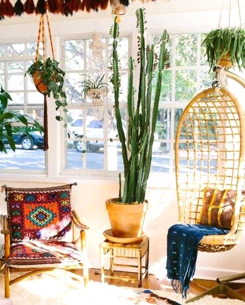 a bright boho sunroom space with a wicker pendant chair, a colorful chair, layered rugs, potted plants and a view