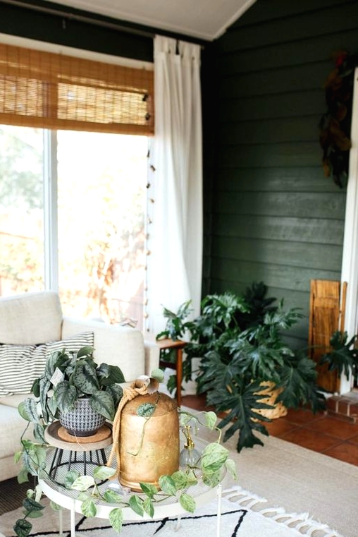 a mid-century modern boho sunroom with green walls, a neutral sectional, round tables, potted plants and woven shades