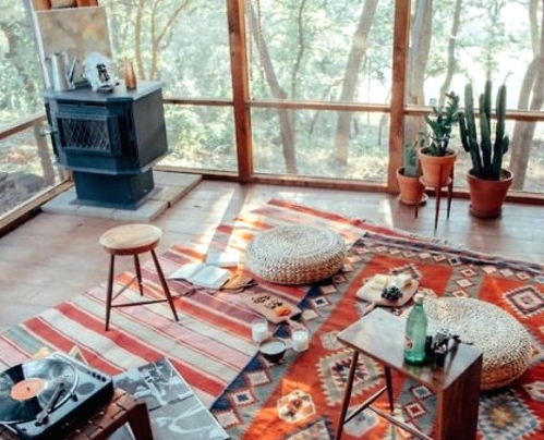 a welcoming boho sunroom with a hearth, colorful boho rugs, jute poufs, potted cacti and stools and a view of the forest