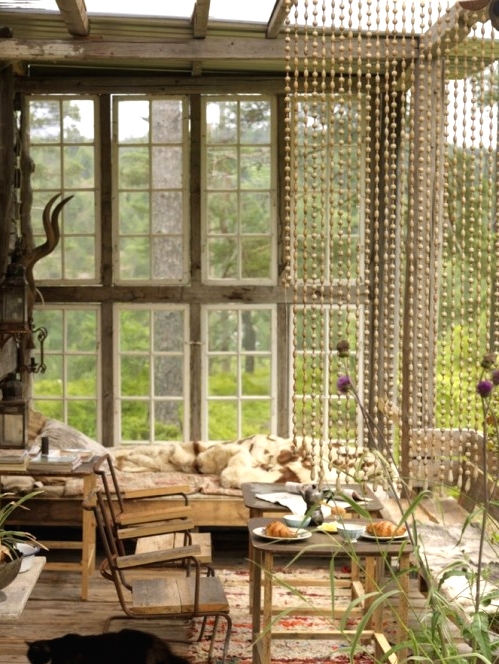 a neutral woodland boho sunroom with a pallet bed and various wooden tables and chairs, wooden beads and antlers, a lovely view of the forest