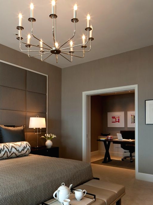 a lovely modern bedroom with taupe walls, a bed with an oversized upholstered headboard, an upholstered bench and a lovely chandelier