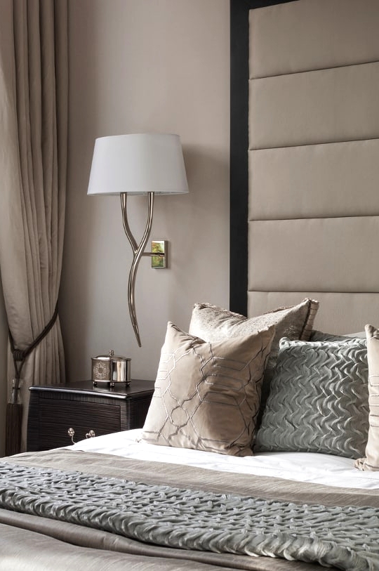 a refined taupe bedroom with a bed with an oversized upholstered headboard, a dark nightstand, a chic sconce and taupe and white bedding