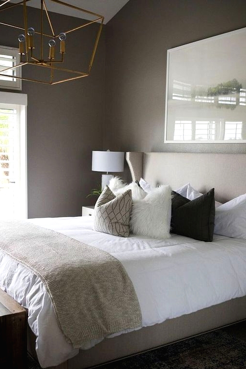 a welcoming taupe bedroom with a tan bed, neutral and printed bedding, a gilded chandelier and a wooden bench