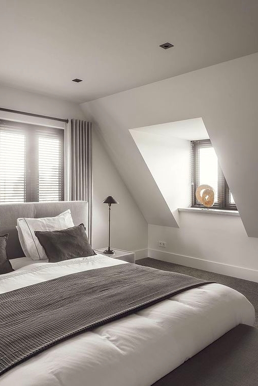 an elegant attic bedroom with windows with dark shades, a grey bed with taupe and white bedding, table lamps