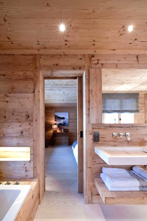 a blonde wood chalet bathroom with a floating sink and vanity, a bathtub clad with stone and a lit up niche