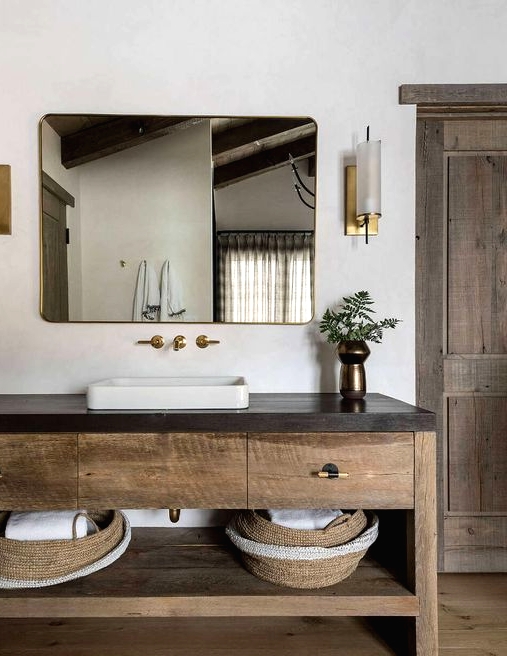 a chalet bathroom done with wood, with a large wooden vanity with a stone countertop, a chic mirror and elegant sconces
