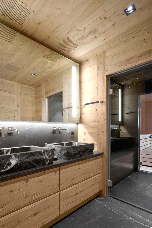 a contemporary chalet bathroom clad with light-stained wood, with stone sinks and a countertop, a lit up mirror and a steam room