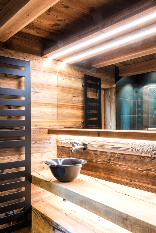 a contemporary chalet bathroom clad with wood, with a lit up shelf vanity, a black bowl sink and a modern radiator