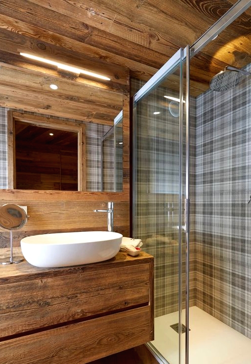 a cool chalet bathroom clad with wood, with a floating vanity, checked tiles in the shower space and a lit up mirror