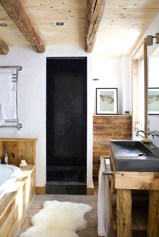 a modern chalet bathroom done with wood, large scale stone tiles, wooden beams on the ceiling, a stone sink and a shower space clad with black tiles