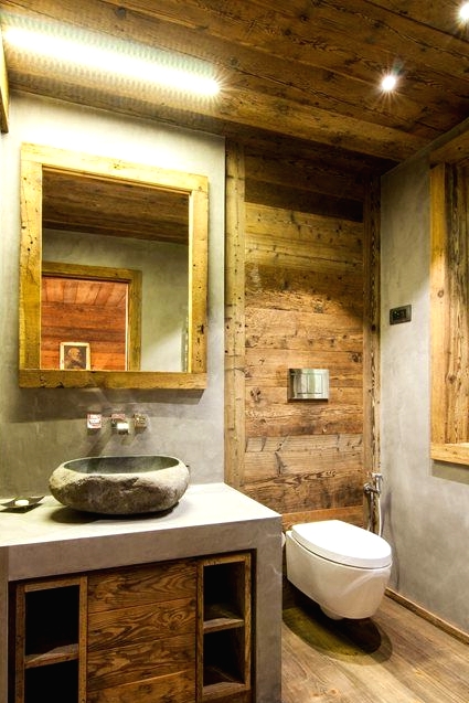 a small chalet bathroom clad with cocnrete and with lots of wood, with a mirror in a wooden frame and a built-in vanity