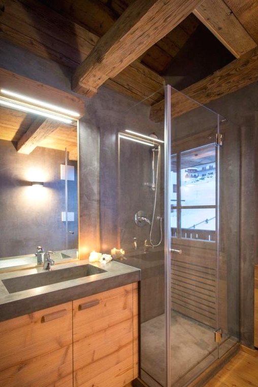 a small minimalist chalet bathroom done with blonde wood and concrete, with textural wooden beams on the ceiling and a lit up mirror