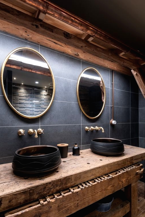 an exquisite chalet bathroom with grey tiles, round mirrors, round black bowl sinks, brass fixtures and a wooden vanity