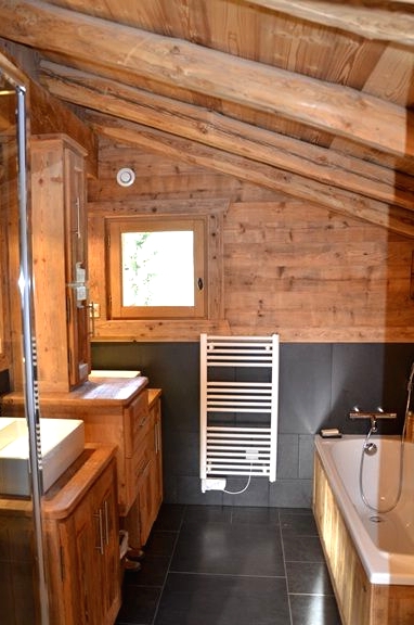 a small chalet bathroom with large scale tiles and light-stained wood, with a small window, wooden furniture and a tub clad with wood