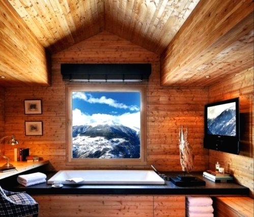 a stylish and cool chalet bathroom clad with light-stained wood, with a view, a TV, a chair, towels, lamps and a blanket