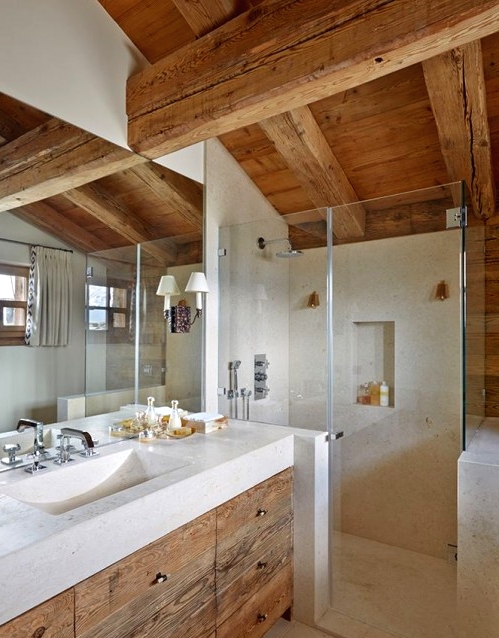a light-filled chalet bathroom with windows, with a light-stained wood ceiling with beams and a matching vanity, neutral stone on the floor, in the shower