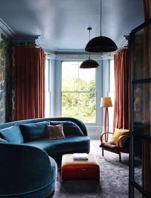 a moody and bold living room with a curved blue sofa, orange textiles and moody bird print wallpaper on one wall